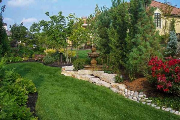  A pineapple shaped water fountain in a mulch bed in the middle of a yard with a background wood line.